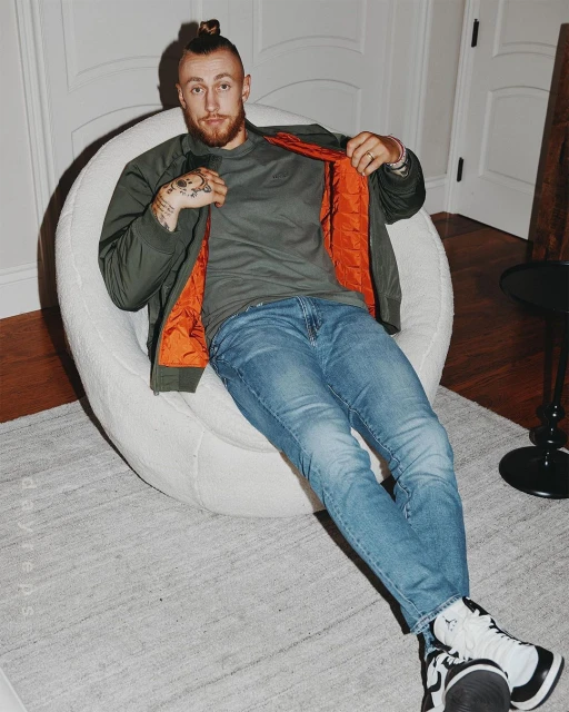 Man in green jacket and jeans sits in a white chair with a light rug, showcasing tattoos and modern style in an indoor setting.