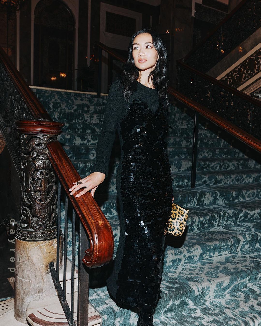 Elegant woman in a long black dress poses on a staircase with patterned carpet and wood railing, holding a leopard print handbag.
