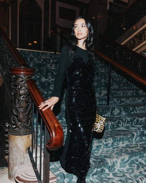 Elegant woman in a long black dress poses on a staircase with patterned carpet and wood railing, holding a leopard print handbag.