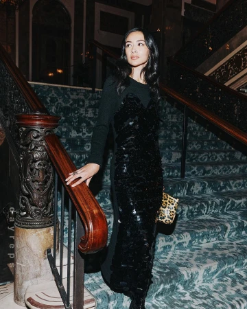 Elegant woman in a long black dress poses on a staircase with patterned carpet and wood railing, holding a leopard print handbag.