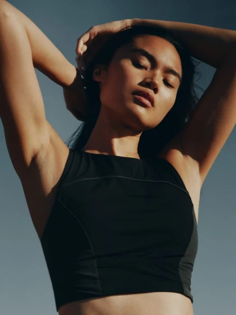 Woman in black sports bra with arms raised against a blue sky. Portrait of a female athlete in fitness apparel.