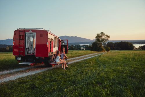 Three people relax on a red camper van parked on a grassy field, with mountains and a lake visible in the distance under a clear sky.