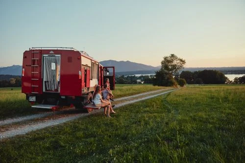 Three people relax on a red camper van parked on a grassy field, with mountains and a lake visible in the distance under a clear sky.