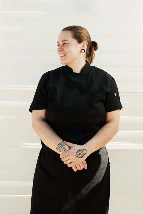 Smiling chef in a black uniform stands on white steps outdoors. The chef has tattoos on their hands and a ponytail.