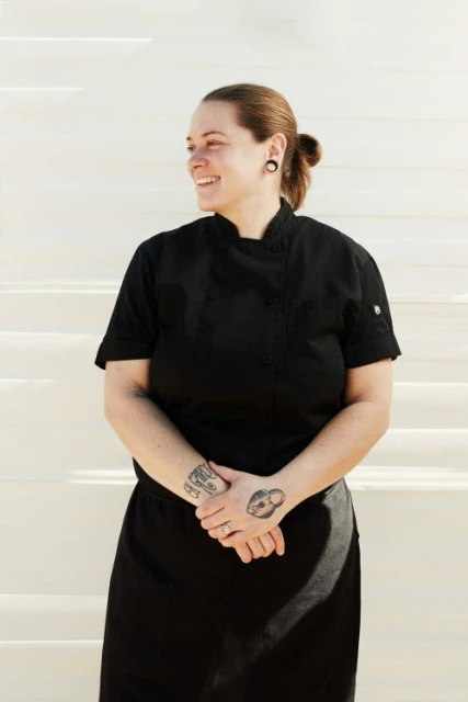 Smiling chef in a black uniform stands on white steps outdoors. The chef has tattoos on their hands and a ponytail.