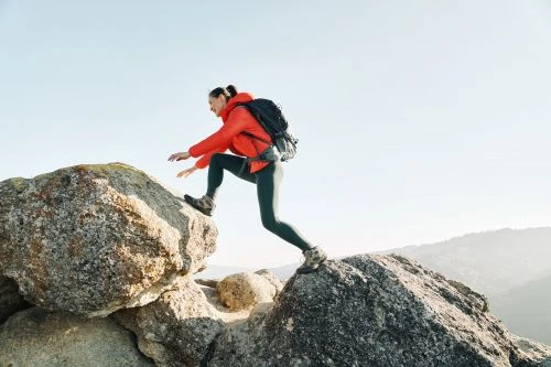 Woman in red jacket climbs rocks outdoors with backpack against clear sky, enjoying a sunny day in a mountainous environment.