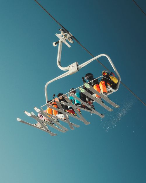 A low-angle view captures a group of people riding up a ski lift on a clear day, showing the mechanics and structure of the lift against the blue sky.