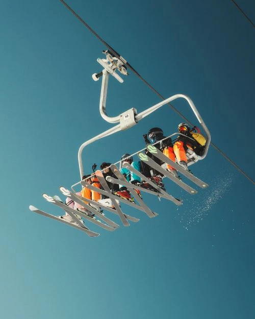 A low-angle view captures a group of people riding up a ski lift on a clear day, showing the mechanics and structure of the lift against the blue sky.