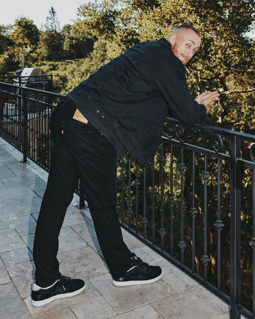Man in black casual wear poses on a balcony with foliage background and neutral stone tiles. Casual outdoor fashion shot.