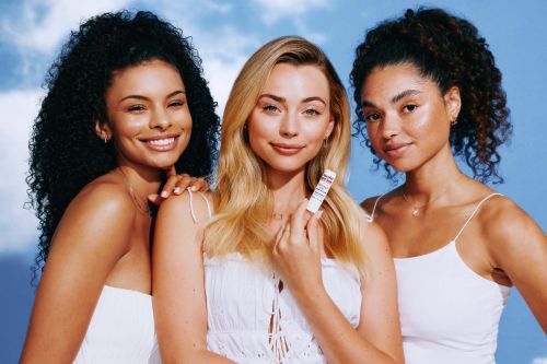 Three diverse women in a beauty shot against a sky backdrop, one holding a skincare product, smiling. Perfect for beauty or skincare campaigns.