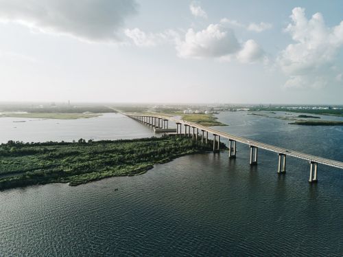 An aerial view captures a long concrete bridge stretching across a wide expanse of dark water, connecting green islands under a partly cloudy sky.