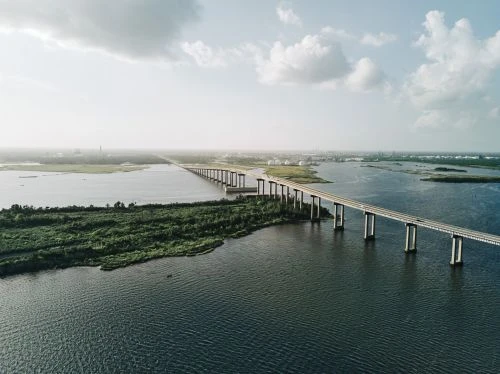 An aerial view captures a long concrete bridge stretching across a wide expanse of dark water, connecting green islands under a partly cloudy sky.