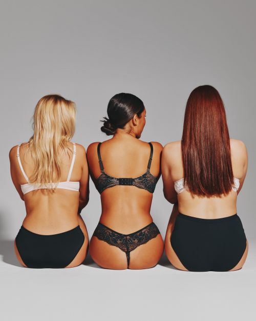 Back view of three diverse adult women in matching lingerie sitting closely together against a neutral grey backdrop in a photo studio.