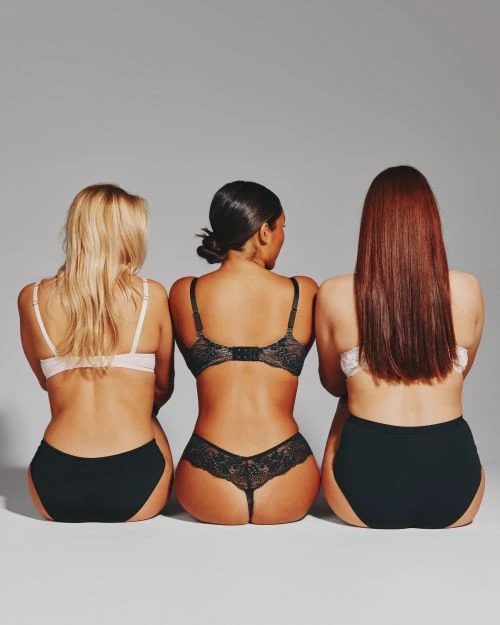 Back view of three diverse adult women in matching lingerie sitting closely together against a neutral grey backdrop in a photo studio.