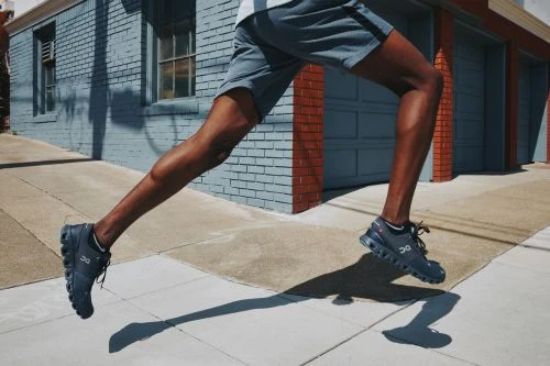 Athletic man mid-stride on a city street, legs and running shoes in sharp focus, blue brick wall and garage doors in the background.