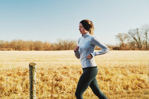 Woman in athletic wear jogs along a rural path in the daytime. She wears a gray long-sleeve shirt and black leggings, near a farm field.