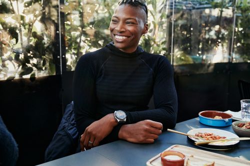Smiling adult woman sits at an outdoor table with snacks and drinks, wearing black top and watch, with a blurred green background.
