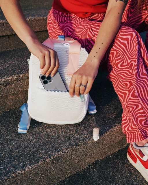 Person holding a white mesh backpack with a visible cell phone in the front pocket, showcasing a casual and youthful style.