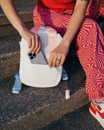 Person holding a white mesh backpack with a visible cell phone in the front pocket, showcasing a casual and youthful style.