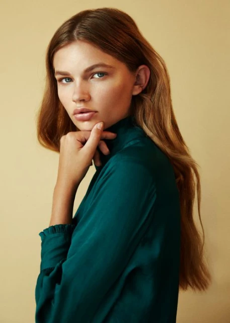 Portrait of a woman with long auburn hair, wearing a dark green top, looking at the camera with a neutral facial expression.
