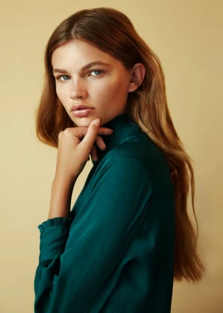 Portrait of a woman with long auburn hair, wearing a dark green top, looking at the camera with a neutral facial expression.