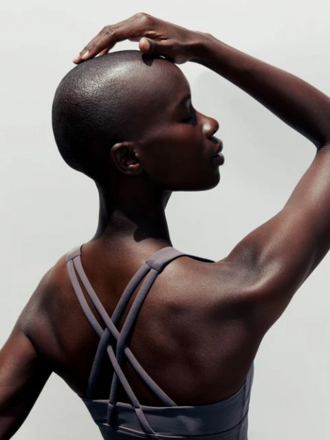 Portrait of a bald African woman with her hand on her head in front of a white background, wearing a gray athletic top.