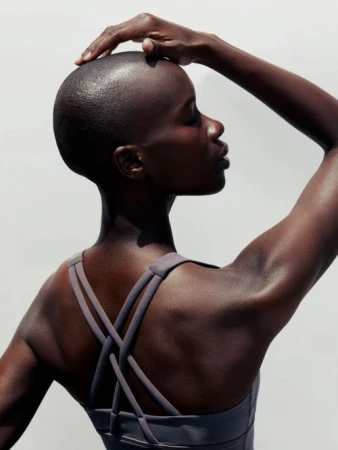 Portrait of a bald African woman with her hand on her head in front of a white background, wearing a gray athletic top.