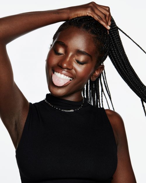 Black woman with long braids wearing a black top holding her ponytail and sticking out her tongue against a white background