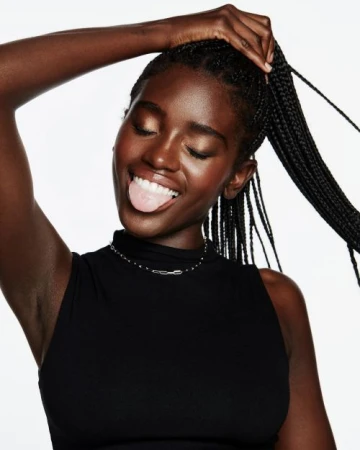 Black woman with long braids wearing a black top holding her ponytail and sticking out her tongue against a white background
