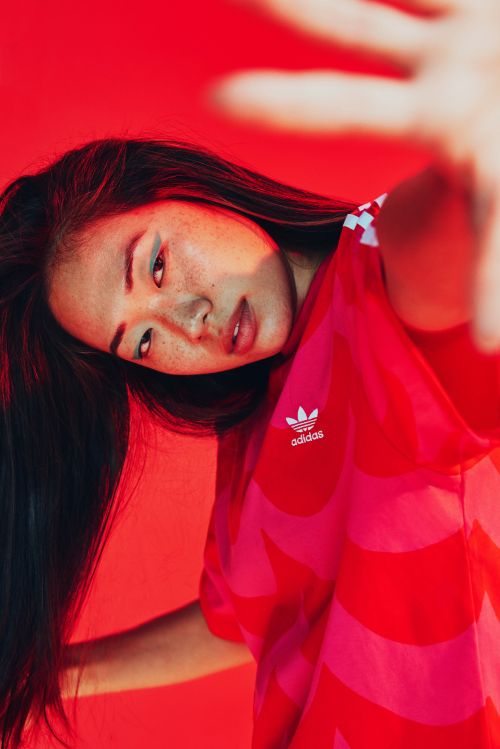 Portrait of a young Asian woman with long dark hair wearing an Adidas shirt, posing in front of a red background with her hand raised.