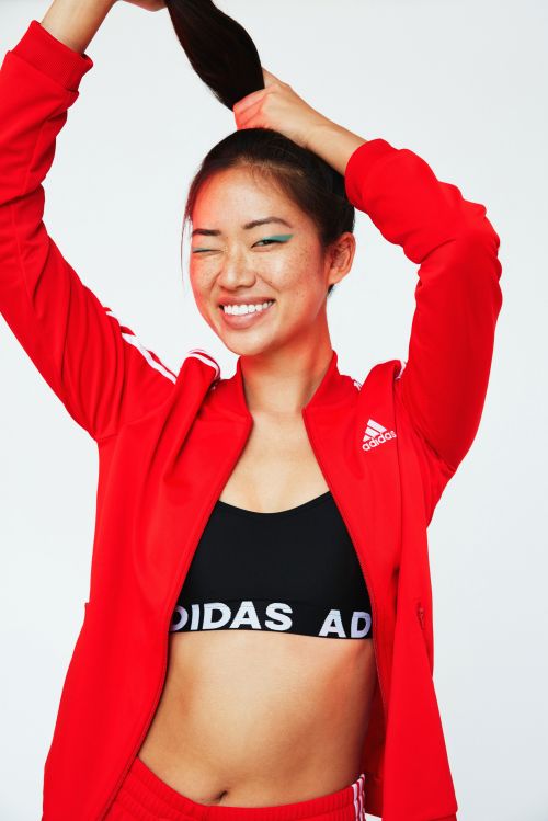 Portrait of a young Asian woman in an Adidas jacket, black top, and shorts, smiling and holding her hair up in a ponytail against a white backdrop.