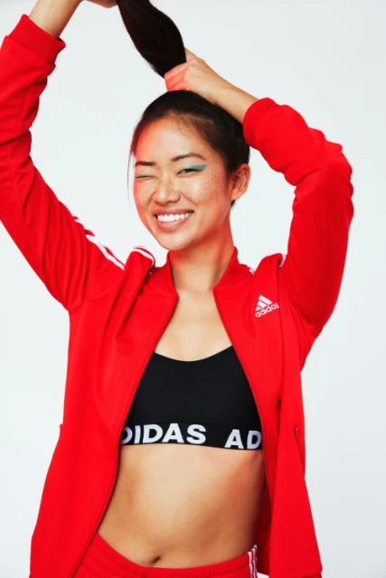 Portrait of a young Asian woman in an Adidas jacket, black top, and shorts, smiling and holding her hair up in a ponytail against a white backdrop.