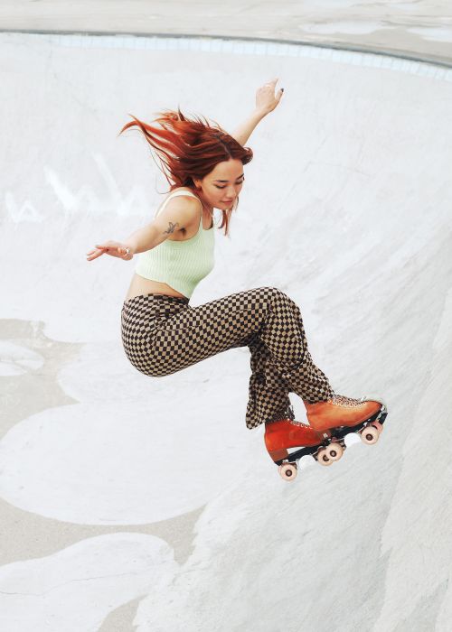 Woman in green top and checkerboard pants mid-air on orange roller skates in a concrete skatepark bowl, arms outstretched.