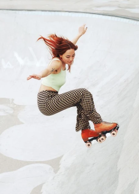 Woman in green top and checkerboard pants mid-air on orange roller skates in a concrete skatepark bowl, arms outstretched.