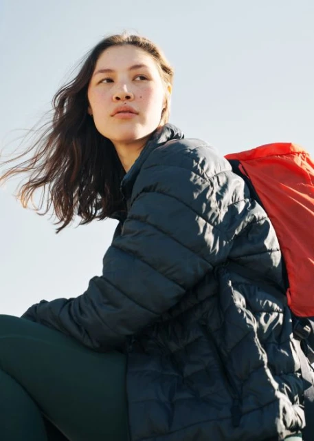 Young woman poses outdoors wearing a black puffer jacket and a red backpack, set against a bright blue sky during daytime.