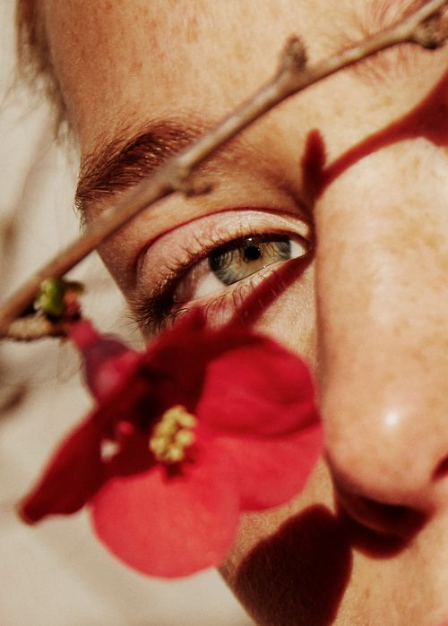 Close-up portrait of a person's eye partially obscured by a delicate twig and red flower by Katie Thompson.