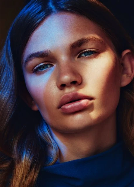 Close-up portrait of a young woman with wavy hair, blue eyes, and fair skin, illuminated by a mix of warm and cool lighting.