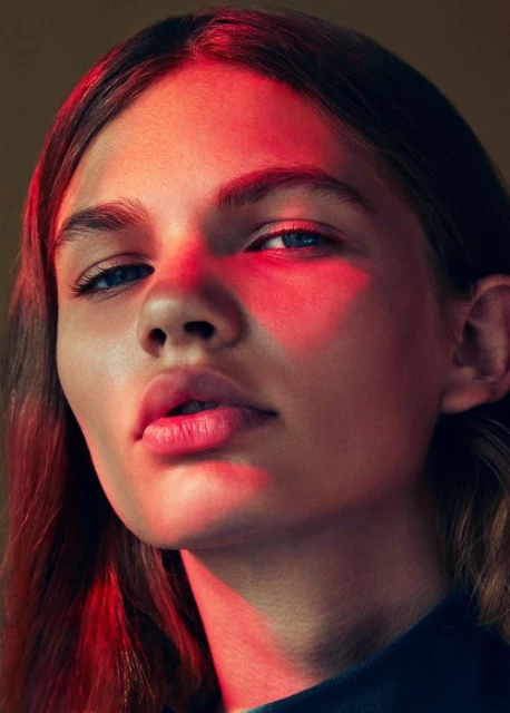 Close-up portrait of a young adult woman with blue eyes, fair skin, and light brown hair, illuminated by dramatic red lighting in a studio setting.