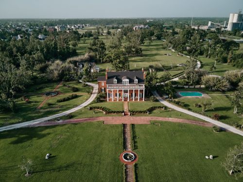 Aerial view of a large brick mansion with white columns on a green lawn, surrounded by trees and a swimming pool on a sunny day.