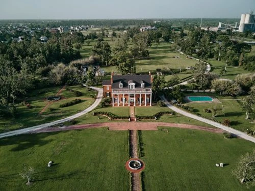 Aerial view of a large brick mansion with white columns on a green lawn, surrounded by trees and a swimming pool on a sunny day.