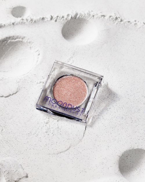 Close-up studio shot of a glittery rose-gold eyeshadow compact in a clear case on a shimmering white surface.