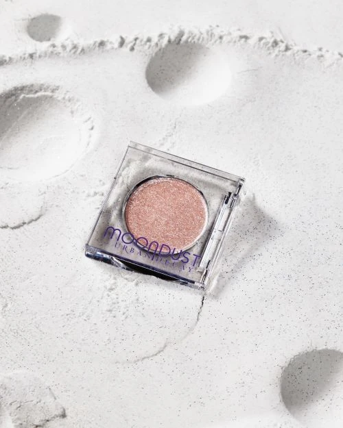 Close-up studio shot of a glittery rose-gold eyeshadow compact in a clear case on a shimmering white surface.