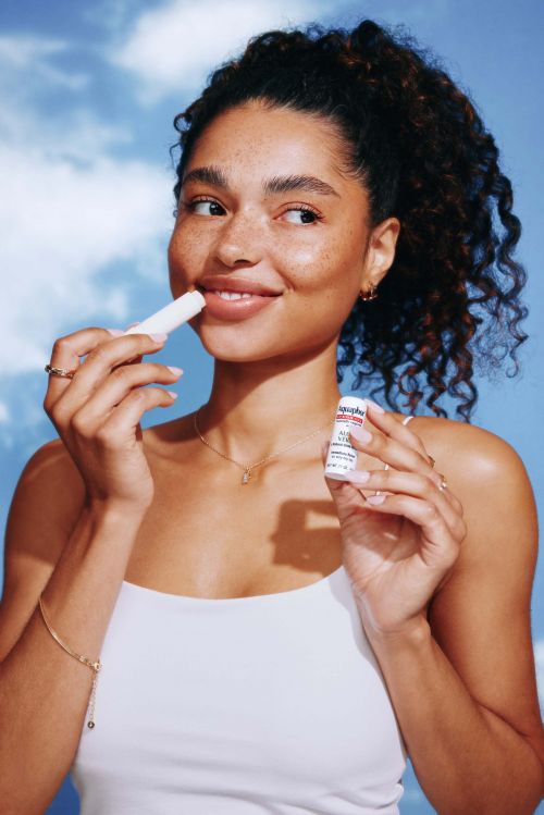 Woman applies lip balm in a bright, sunny scene. The focus is on the lip care product and the model's natural beauty.