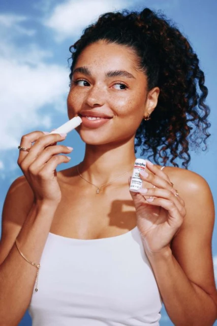 Woman applies lip balm in a bright, sunny scene. The focus is on the lip care product and the model's natural beauty.