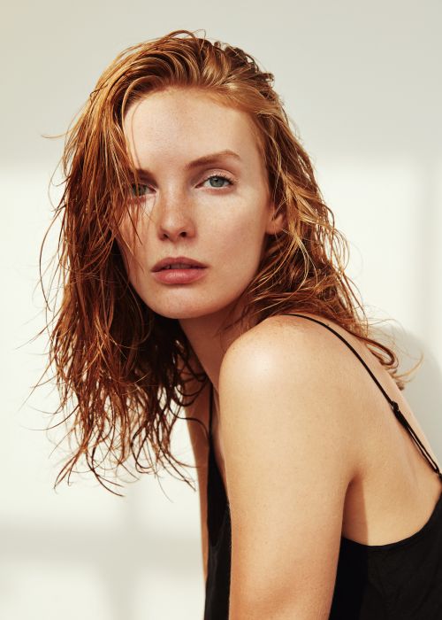 Portrait of a woman with wet ginger hair and freckles wearing a black strap top against a white wall.