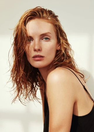 Portrait of a woman with wet ginger hair and freckles wearing a black strap top against a white wall.