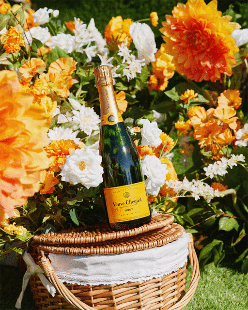 Veuve Clicquot champagne bottle on wicker picnic basket surrounded by white and orange flowers and green foliage.