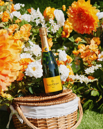 Veuve Clicquot champagne bottle on wicker picnic basket surrounded by white and orange flowers and green foliage.