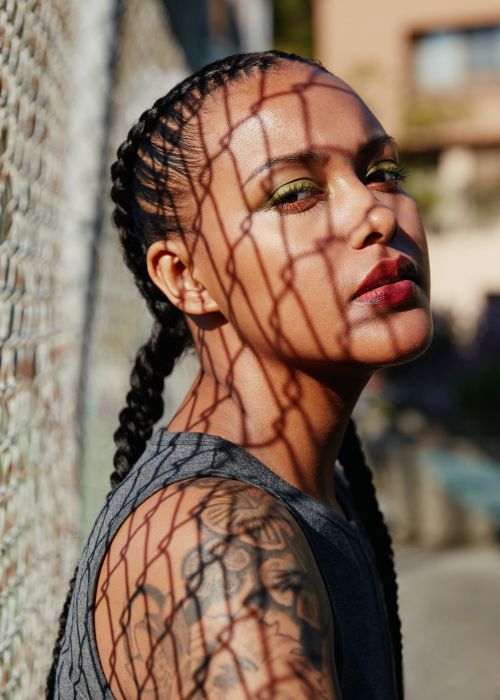 Portrait of a woman with braided hair and olive eyeshadow, posing in front of a fence creating shadow patterns on her face.