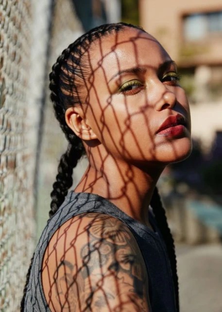Portrait of a woman with braided hair and olive eyeshadow, posing in front of a fence creating shadow patterns on her face.